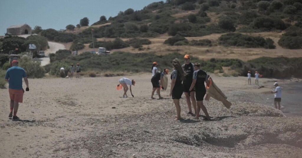 Εθελοντές καθαρίζουν την παραλία της Αναβύσσου στο πλαίσιο δράσης του Saronikos Synergy σε συνεργασία με τη Betsson. Άτομα με πορτοκαλί καπέλα και σακούλες σκουπιδιών συλλέγουν απορρίμματα στην ακτή, συμβάλλοντας στην προστασία του περιβάλλοντος και την ευαισθητοποίηση της τοπικής κοινωνίας.